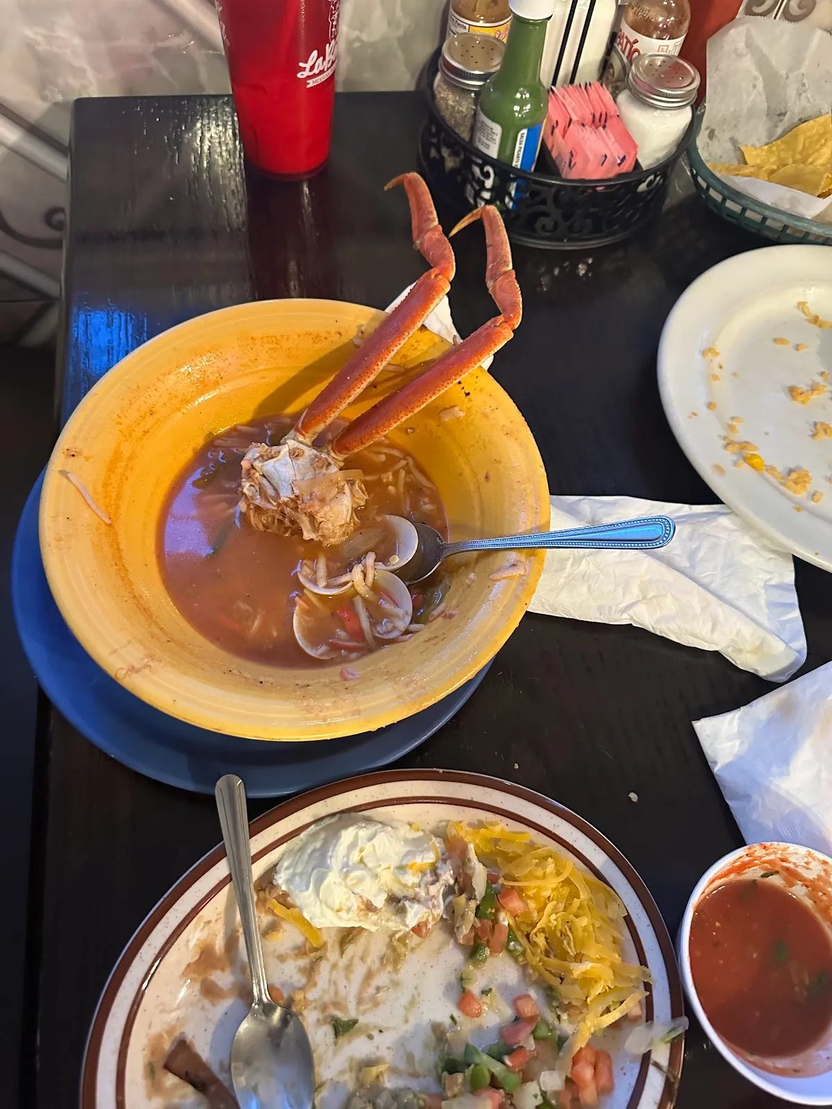 A bowl of seafood caldo with a crab leg in a yellow bowl, served with tortilla chips and salsa