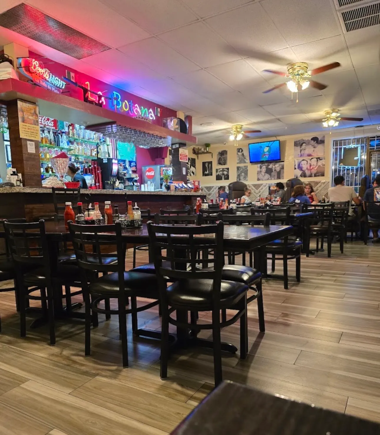 La Botana's dining room with wood floors, black tables, family photos on the wall, and the La Botana sign lit over the bar