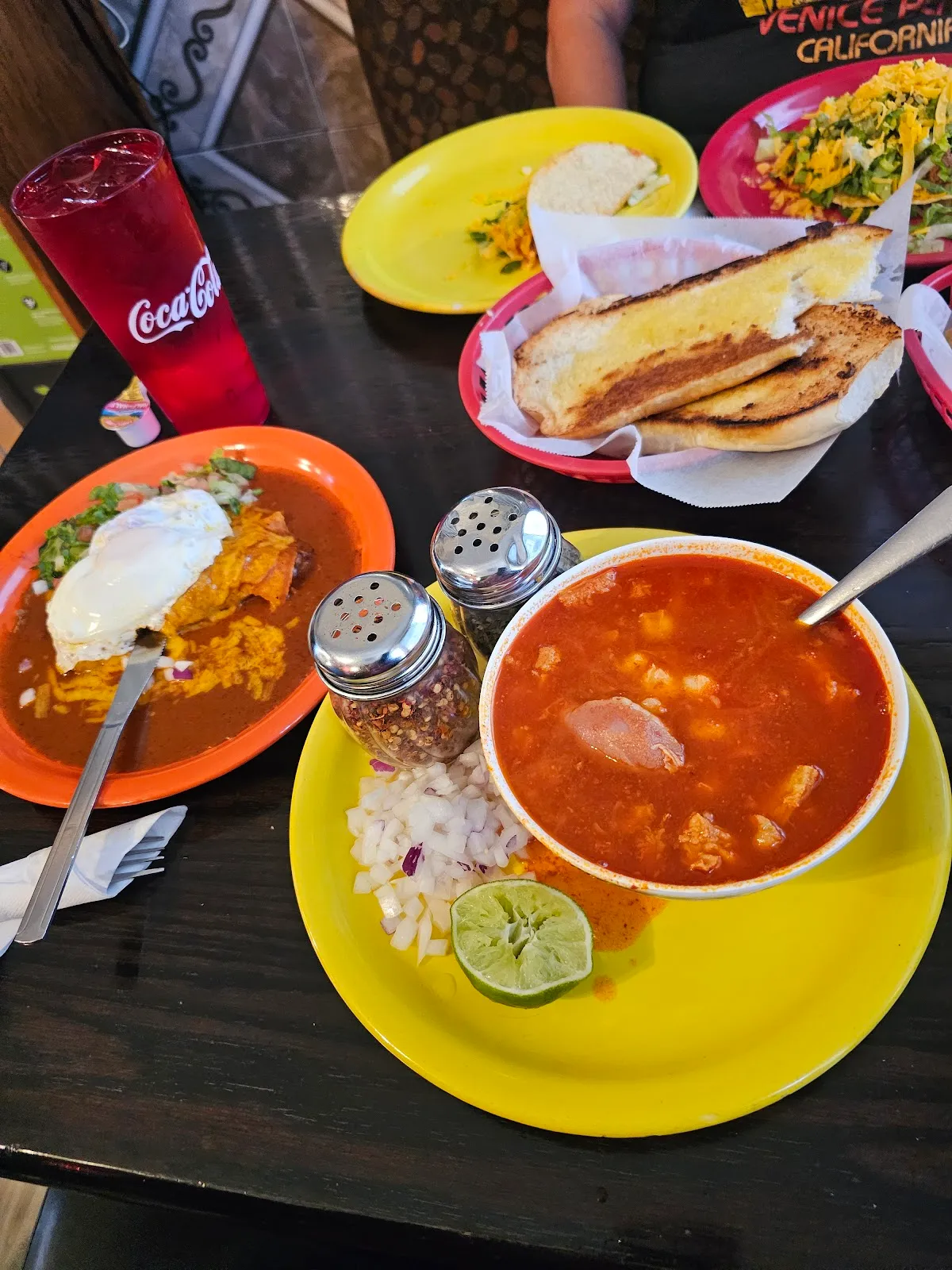 A table spread with a bowl of red soup, enchiladas topped with a fried egg, a basket of toasted bread, and a Coca-Cola