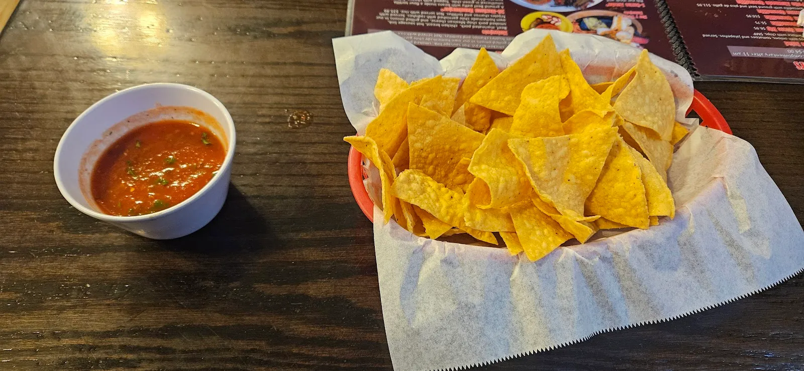A basket of warm tortilla chips next to a cup of red salsa on a dark wood table